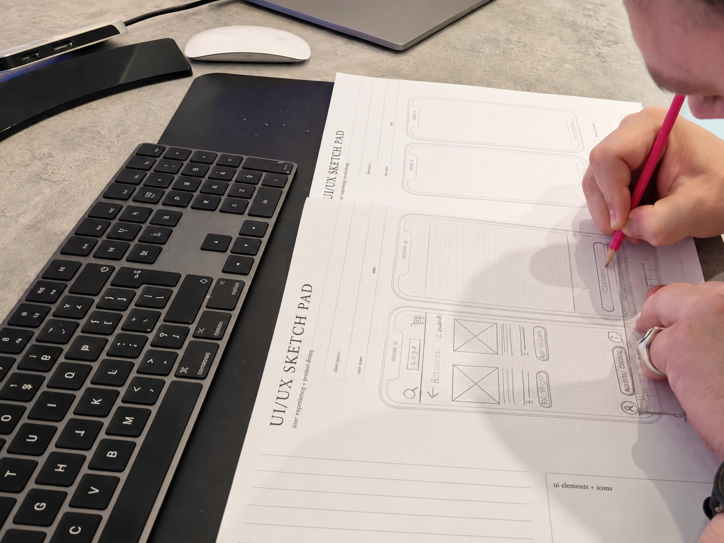 Designer sketching mobile app screens on a UI/UX sketch pad next to a keyboard